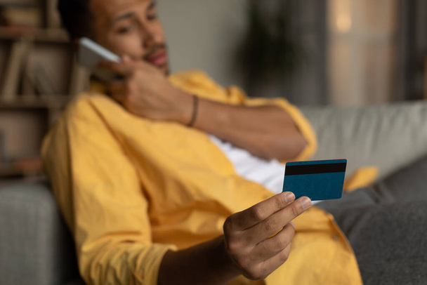 Young African American man holding credit card, calling to bank client service, having problem during online shopping at home, selective focus. Remote banking, electronic money concept - Foto, imagen