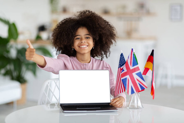 Happy black girl with bushy hair schooler sitting at desk with international flags, showing computer with white empty screen and thumb up, child learning foreign languages online, mockup, copy space - Фото, изображение