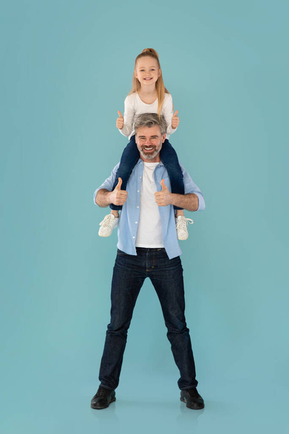 Cheerful Father Holding Daughter On Shoulders Gesturing Thumbs Up Approving Offer Together On Blue Studio Background. We Like And Approve It Concept. Vertical, Full Length Shot - Foto, immagini