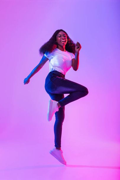 Joyful millennial African American woman flying in air, jumping, smiling at camera in neon light, full length portrait. Expressing positivity, joy and happiness concept - 写真・画像