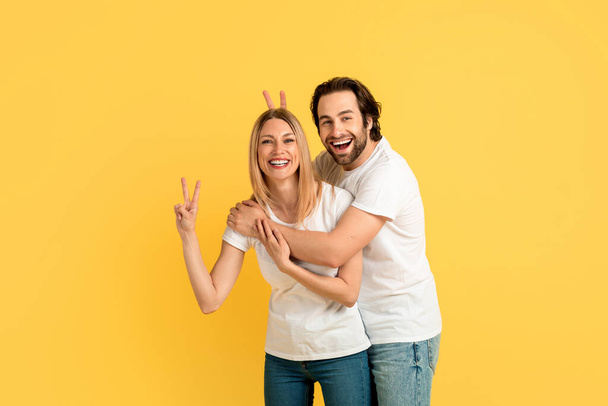 Cheerful funny millennial caucasian woman and man in white t-shirts have fun, show peace sign with hands, isolated on yellow background, studio. Couple enjoy free time, love and family relationship - Valokuva, kuva