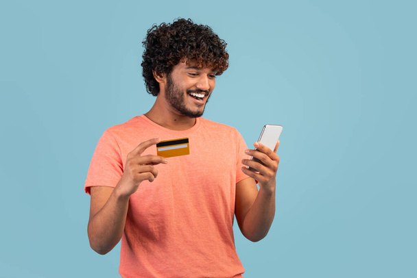 Happy millennial indian guy with curly hair in pink t-shirt holding modern cell phone and yellow credit card, shopping or banking online, using mobile application, blue studio background - Foto, Bild