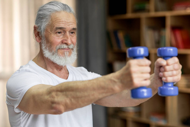 Athletic grey-haired bearded old man in sportswear exercising alone at home, using blue barbells, working on arms strength, pumping iron, closeup photo, side view, copy space - Foto, Imagem