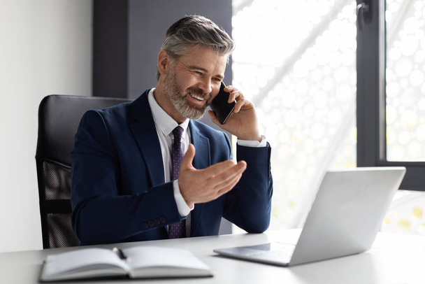 Corporate Communication. Handsome Middle Eaged Businessman Talking On Cellphone In Office, Smiling Male Entrepreneur In Suit Having Mobile Conversation While Sitting At Desk With Laptop, Copy Space - Фото, зображення