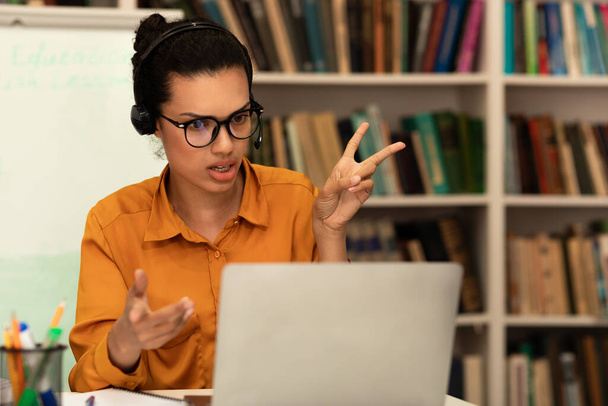 Remote distant communication concept. Mixed race female teacher in headphones teaching foreign languages, talking and gesturing at laptop webcamera during online call, sitting in library - Foto, imagen