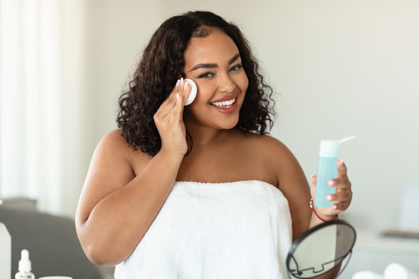 Beautiful black chubby lady cleaning her face, using cotton pads and cleansing product, looking at camera and smiling. Young lady using face toner and cotton pad, home interior - Foto, Imagen
