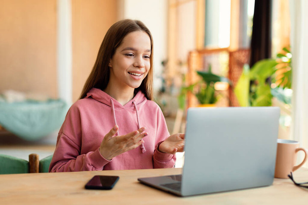 Positive teen girl video calling on laptop and gesturing at webcamera, sitting at desk at home. Cheerful teenager chatting with friends online by social distance virtual videocall - Fotoğraf, Görsel
