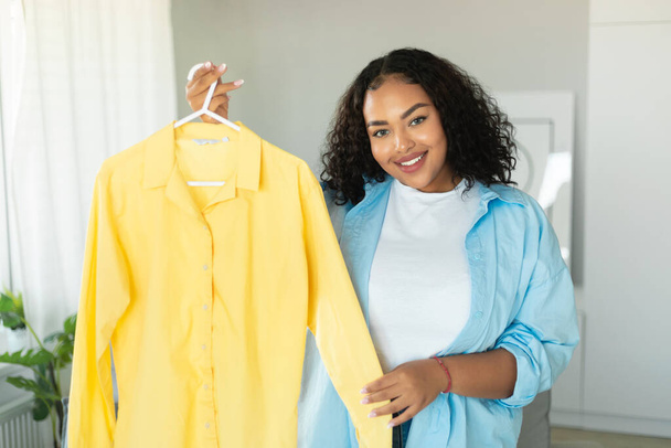 Шопінг і стиль. Щаслива чорна жінка вішалка для шопоголіка з сорочкою, що показує новий одяг, що стоїть вдома, посміхається камері. Концепція моди та споживачів
 - Фото, зображення