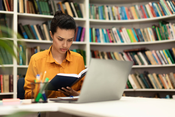 Concentrated mixed race woman reading book, sitting in library with laptop pc and study materials, free copy space. Remote education, distance learning concept - Foto, Imagem