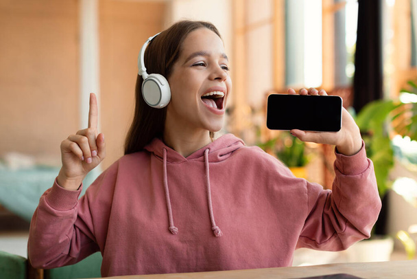 Joyful teen girl singing song and using smartphone as microphone, wearing wireless headset, having fun at home. Carefree teenager enjoying free time after online classes - Photo, Image