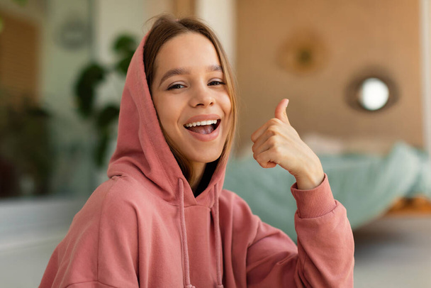 I like it. Cheerful stylish teen girl showing thumbs up gesture, approving or recommending something, sitting at home and smiling at camera. Positive adolescent expressing her agreement - Foto, immagini