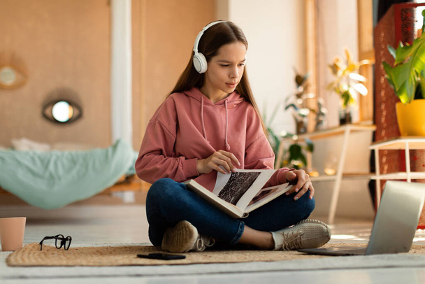Focused caucasian teen girl sitting in wireless headphones and holding book, looking at laptop screen in bedroom interior at home, copy space - Zdjęcie, obraz