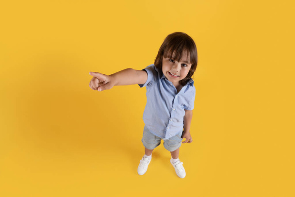 Look here. Top view portrait of cute little boy pointing aside at free space and smiling, yellow studio background - Zdjęcie, obraz