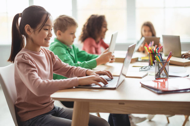Електронне навчання. Азіатська маленька дівчинка використовує ноутбук комп'ютер сидячи з різними однокласниками в сучасному класі в школі. Технології та сучасна освіта. Вибірковий фокус
 - Фото, зображення