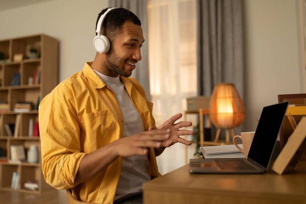 Happy young black man using laptop, wearing headphones, having video chat at home, copy space. African American male communicating online, working or studying remotely on portable pc - 写真・画像