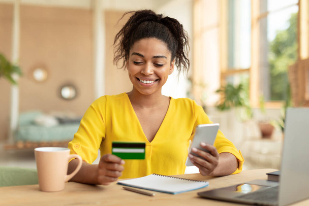 Positive african american lady with smartphone and credit card purchasing goods or services on web, sitting at desk at home. Happy black woman buying products online - Foto, immagini