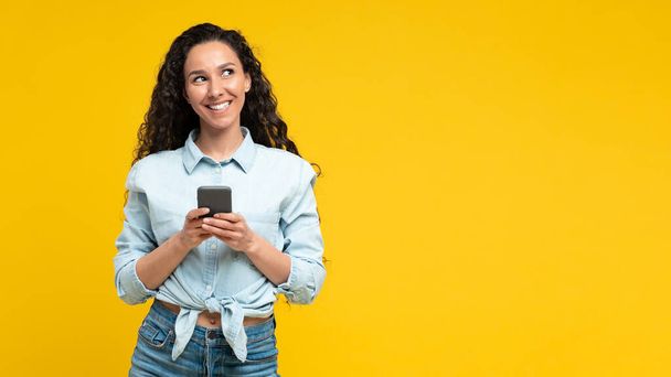 Cheerful Arabic Woman Using Mobile Phone Browsing Internet And Texting Standing On Yellow Background. New Application Advertisement Concept. Panorama, Studio Shot - 写真・画像