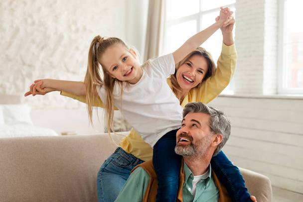 Joyful Family Of Three Posing And Having Fun, Kid Daughter Sitting On Dads Shoulders Bonding At Home. Middle Aged Parents And Child Playing Together On Weekend - Photo, Image