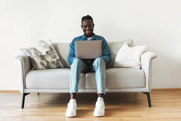 Чорний фрілансер хлопець використовує ноутбук комп'ютер працює онлайн сидячи на дивані вдома. Щасливий хлопець Websurfing в сучасній вітальні в приміщенні. Велика концепція реклами сайту
 - Фото, зображення