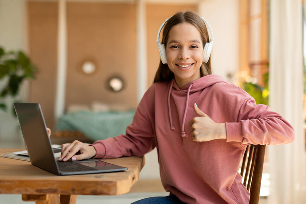 Happy teen schoolgirl in headset gesturing thumb up, sitting near laptop, having online lesson from home. Cheerful teenager expressing approval, recommending online school or educational course - Photo, Image