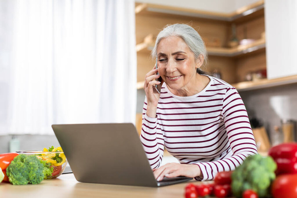 Beautiful senior woman talking on cellphone and using laptop in kitchen, smiling elderly freelancer lady working on computer or making purchase online and having mobile conversation, copy space - Photo, image