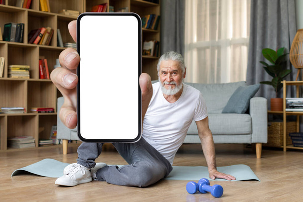 Cheerful handsome athletic senior man sitting on yoga mat, showing modern cell phone with white empty screen, recommending nice fitness mobile application, home interior, copy space, mockup - Photo, image