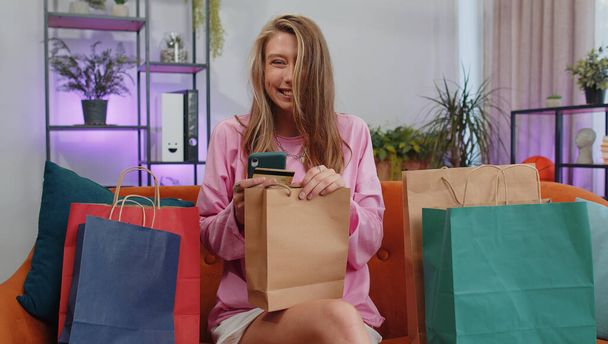 Happy shopaholic consumer girl sitting with shopping bags at modern home apartment making online order, payment with credit plastic bank card. Young woman on couch at home. Discount sale on internet - Valokuva, kuva