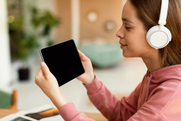 E-learning concept. Positive schoolgirl using digital tablet with empty screen, watching online lesson or surfing internet, wearing headphones. Mockup for your design - Foto, imagen