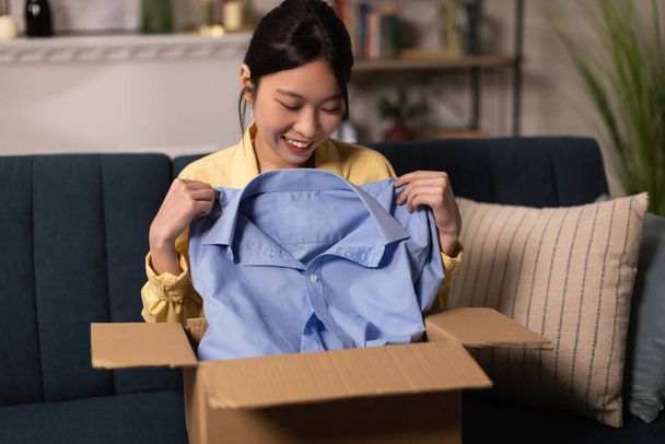 Shopping And Delivery. Joyful Chinese Lady Unpacking Cardboard Box Holding Blue Shirt Receiving Parcel From Shop Sitting On Couch At Home. Ecommerce Service, Contented Customer Concept - Foto, Imagen