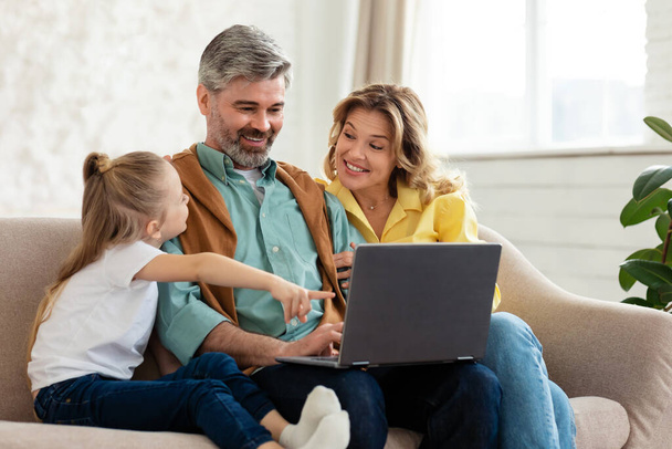 Cheerful Family Of Three Using Laptop Computer, Kid Daughter Pointing Finger Browsing Internet And Recommending Website Sitting On Sofa At Home. Technology And Domestic Lifestyle Concept - Fotoğraf, Görsel