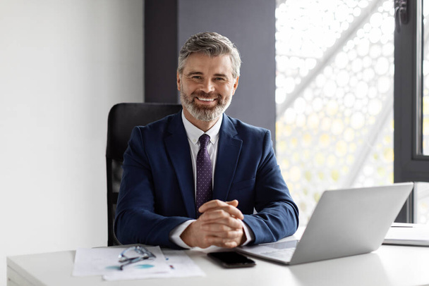 Portrait Of Handsome Smiling Middle Aged Businessman Sitting At Workplace In Office And Looking At Camera, Successful Male Entrepreneur Working At Desk With Laptop Computer, Copy Space - 写真・画像