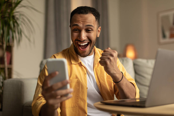 Excited millennial black man with laptop looking at smartphone screen, celebrating online win or work success at home. Young African American guy gambling on internet, enjoying huge sale in web store - Foto, Imagen