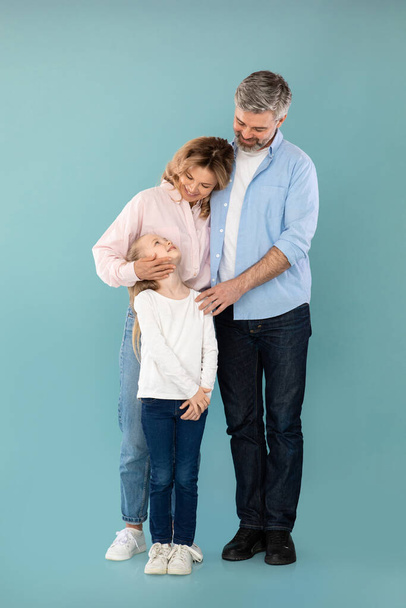 Vertical Shot Of Cheerful Family Embracing Expressing Love Posing Together Over Blue Background In Studio. Mature Parents Hugging Standing With Kid Daughter. Full Length - Fotoğraf, Görsel