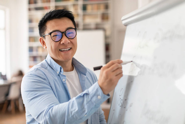 Cheerful Chinese Tutor Smiling To Camera Teaching English Language Writing Grammar Rules On Whiteboard Standing In Classroom Indoor. Modern School Education Concept - 写真・画像