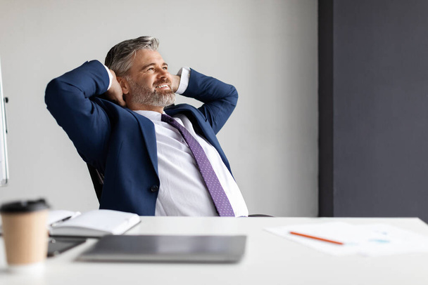 Smiling middle aged businessman leaning back in chair at workplace in office, handsome happy male entrepreneur in suit sitting at desk with hands behind head, resting after successful day - Foto, afbeelding
