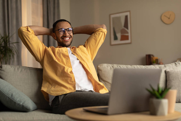 Cheery young black man with laptop relaxing after online job, sitting on sofa with laptop at home, empty space. Cool millennial guy having break while working remotely on web - Valokuva, kuva