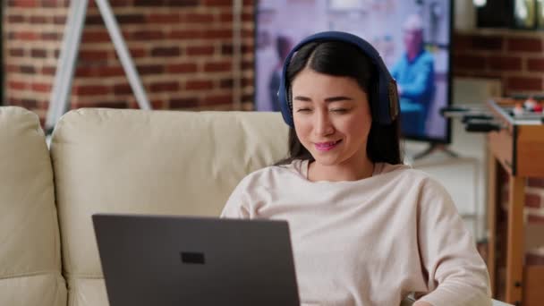 Happy young adult person watching online streaming series of favourite internet show while sitting on sofa at home. Asian woman enjoying watching entertaining comedy movies and content on laptop. - Materiał filmowy, wideo