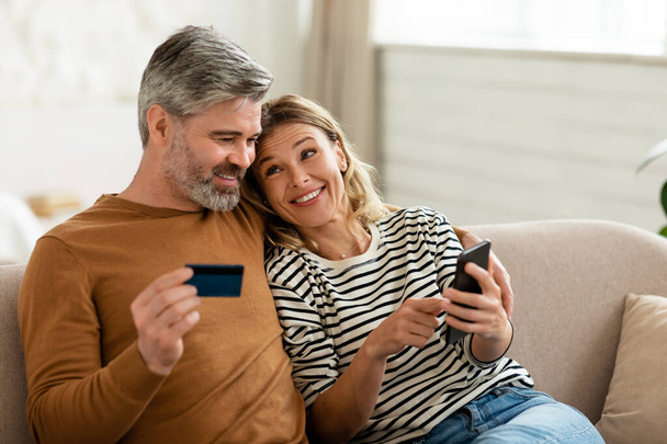 Happy Spouses Shopping Online Via Phone Holding Credit Card Sitting On Sofa At Home. Couple Paying Via Banking Application On Smartphone. Selective Focus - Foto, immagini