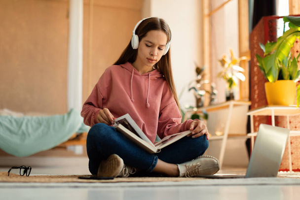 Full length portrait of focused teen girl with book and laptop having lesson, attending online class from home. Female teenage student studying remotely - Valokuva, kuva