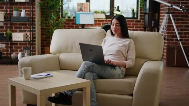 Focused woman sitting on sofa while working remotely on laptop. Beautiful young adult asian student attending online class while typing important lessons on modern portable computer at home. - Video