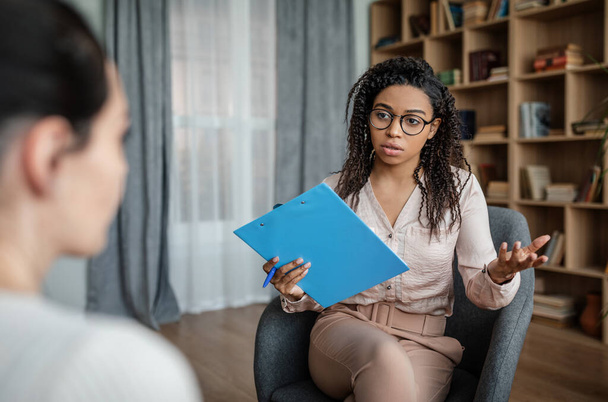Serious african american young woman psychologist advises european lady patient in clinic interior. Mental problems, professional medical care, fight against depression, work with client due covid-19 - Photo, image