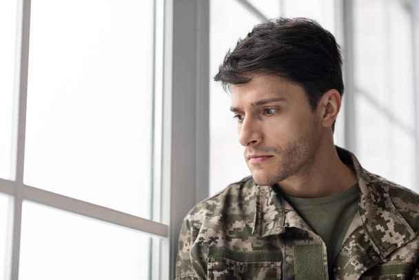 Serious short-haired young man in military uniform soldier looking through the window, upset soldier suffering from posttraumatic syndrome after returning home, copy space, closeup - Photo, image