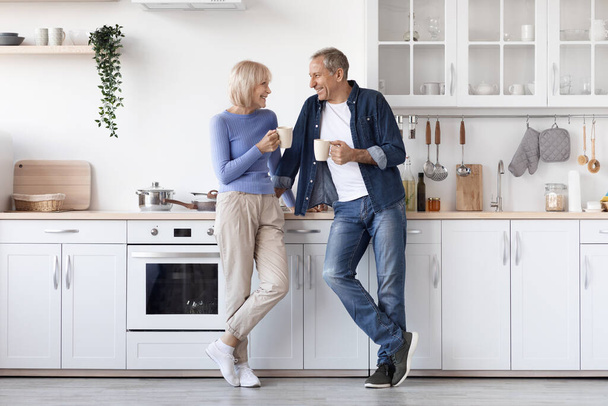 Happy attractive senior couple in homewear standing at white cozy kitchen, drinking fresh morning coffee together, enjoying good morning, sharing plans for day, panorama, copy space, full length - Foto, immagini