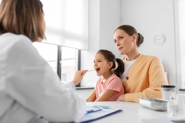 medicine, healthcare and pediatry concept - mother with little daughter and doctor with tongue depressor checking little girl patients throat at clinic - Foto, Bild