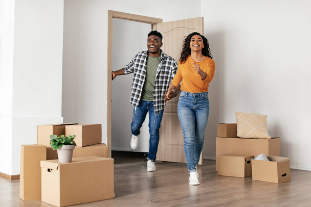 Real Estate Buyers. Joyful Black Couple Entering New Rented Apartment Holding Hands Posing Among Moving Boxes At Home. Property Ownership, Rent And Purchase Concept - Photo, image