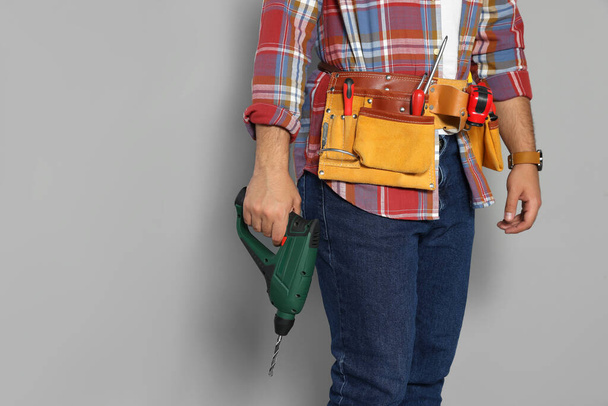 Worker with power drill and tool belt on grey background, closeup. Space for text - Foto, afbeelding