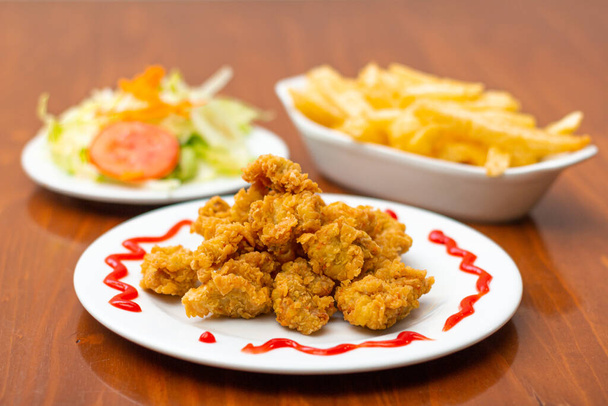 Chicken chicharron, accompanied by French fries and salad on a wooden table. - Фото, зображення
