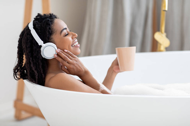 Beautiful Young Black Lady Taking Bath, Listening Music And Drinking Coffee, Smiling African American Woman Wearing Wireless Headphones, Enjoying Relaxing In Bathroom At Home, Side View, Copy Space - Foto, afbeelding