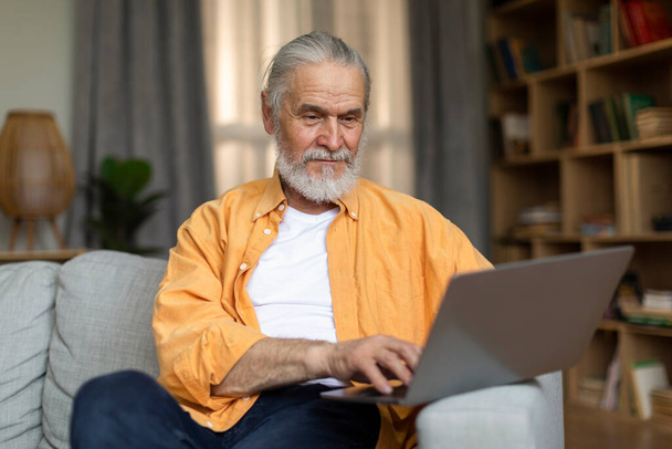 Handsome stylish grey-haired senior man websurfing while resting at home, sitting on couch and using modern computer, living room interior, looking at notebook screen and smiling, copy space - Photo, Image