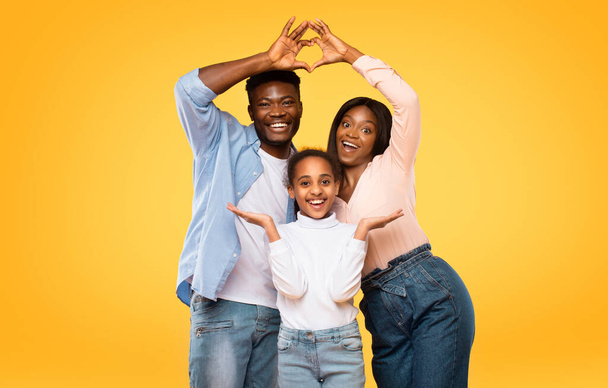 Love, care and support. Excited african american parents showing heart shape with fingers, making symbolic roof under their daughter head over yellow studio background - Zdjęcie, obraz